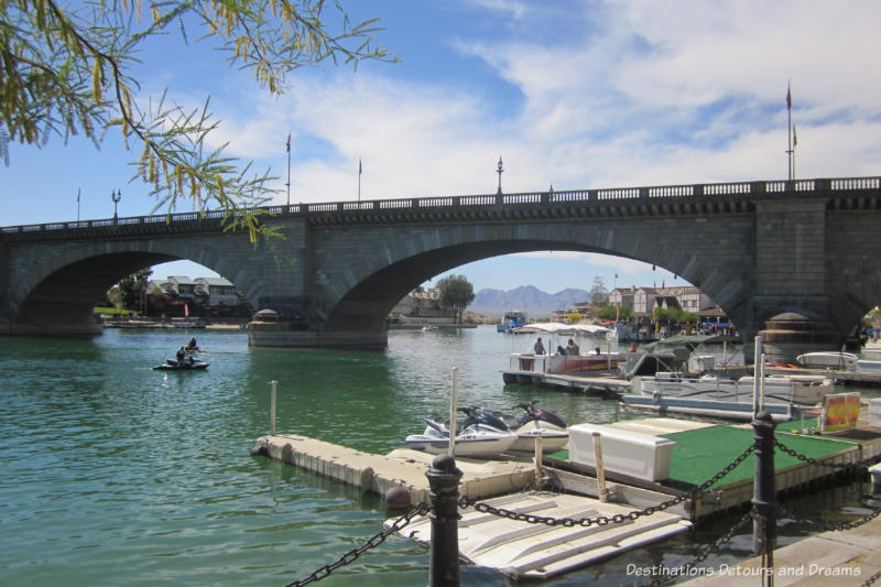 Stone arched bridge over water with boats moored at water edge and mountains in background