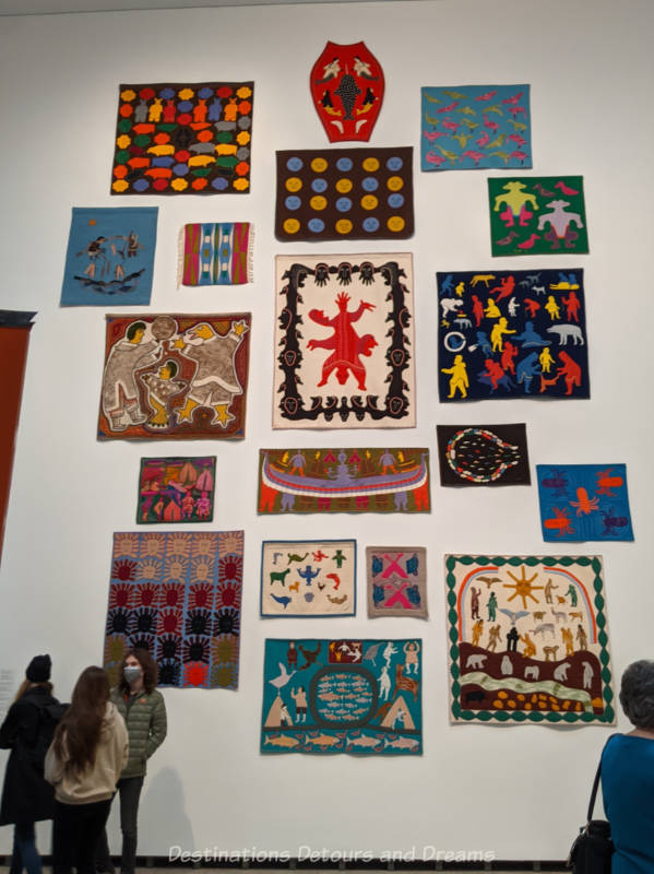Collection of colourful Inuit woolen wall hangings with hand-stitched figures displayed on a two-story white wall at Qaumajuq