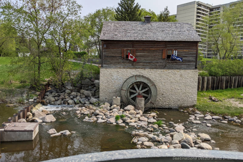 The side of water-powered mill along a creek with stone foundation, weathered wood buildings, and large water wheel.