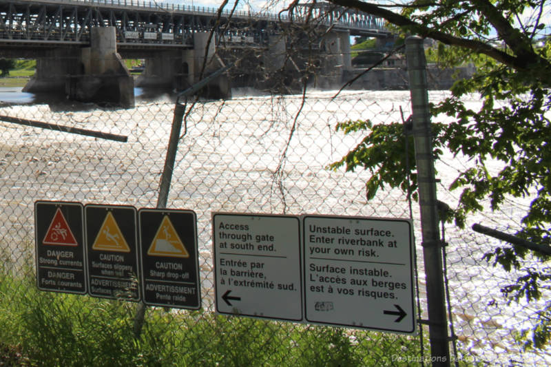 Fencing with view of bridge, dam, and locks in background and signs on fence about entry