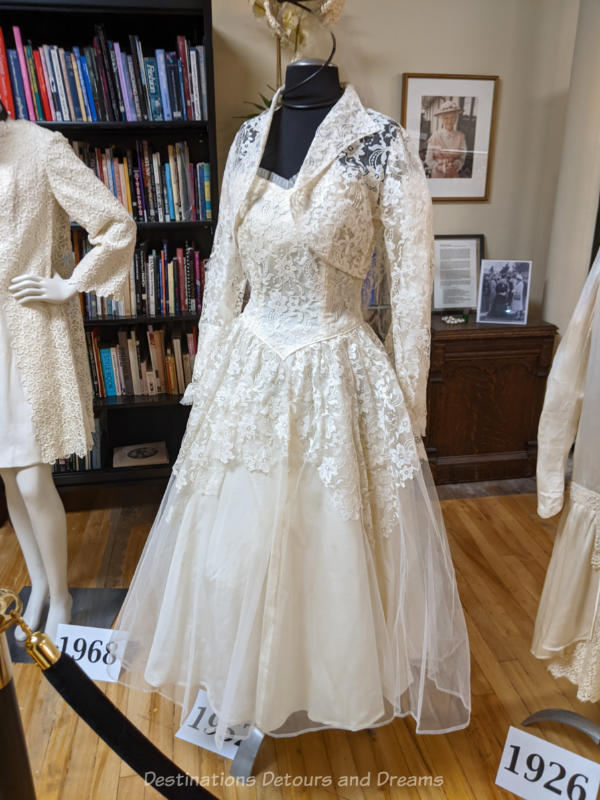 A 1950s white wedding dress with lace bodice and top skirt, v-neck, and a bolero-style lace jacket on a headless mannequin in a museum display