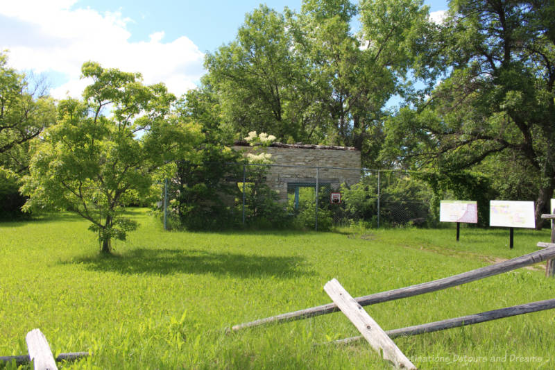 Ruins of an 1850s stone house behind fences on a grassy and treed lot with a few signs about the historic site