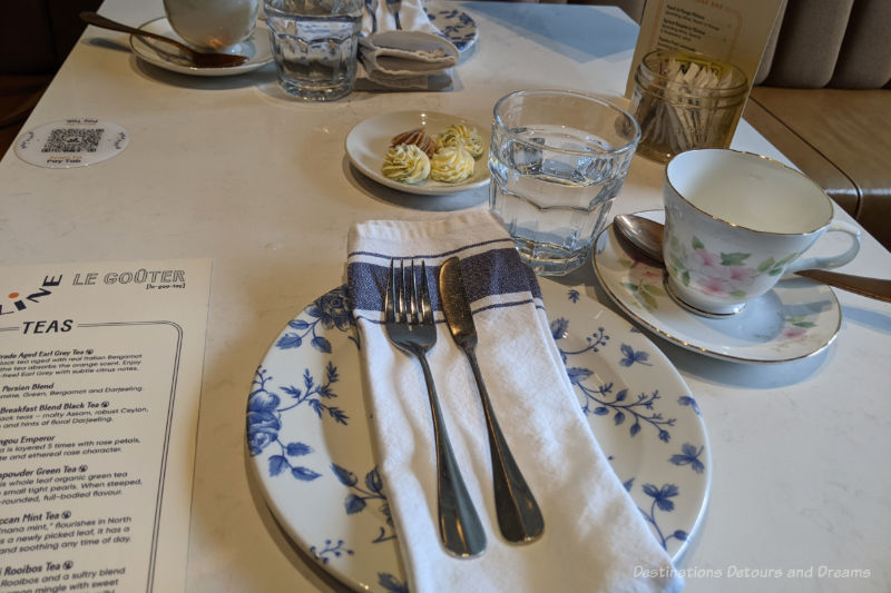 Table setting with white tablecloth, china plate containing napkin, fork, and butter knife, glass of water, and china tea cup ready for French High Tea