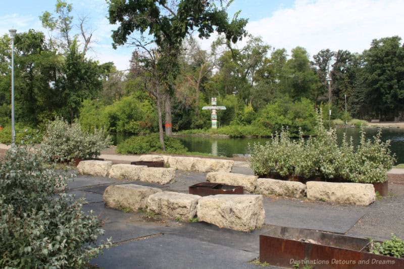 colourful totem pole on mini-island in pond as viewed from a plaza area beside the pond with stone seating