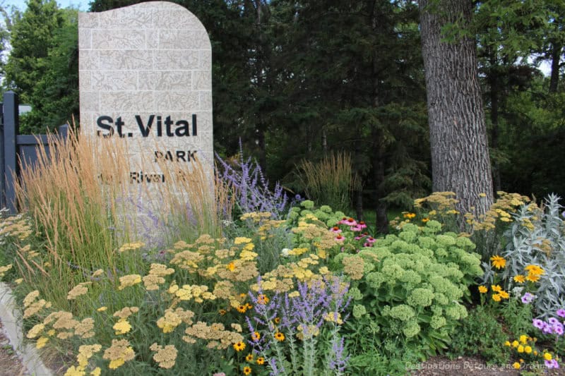 Stone signage amid flower bed in front of trees at entrance to St. Vital Park