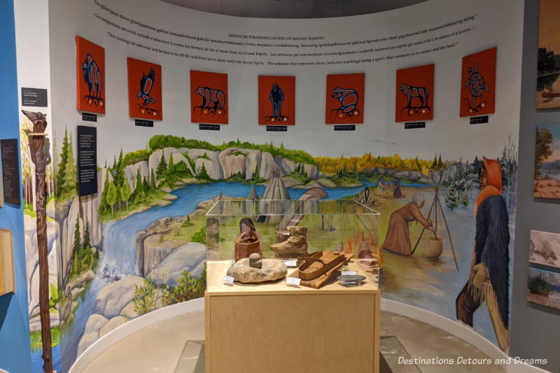 Museum display with glass display case on Indigenous life objects in front of a curved wall contains a mural and panels with the Seven Grandfather Teachings