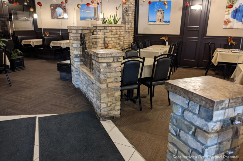 Interior of a restaurant with brick divider walls, tables covered with beige tablecloths, black chairs and pictures of Greece on the walls