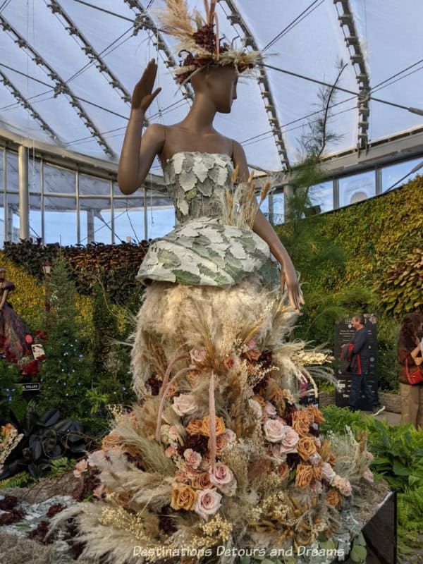 Floral art display featuring mannequin in dress of white and pale green leaves, with skirt of wheat and other flowers in a homage to the architecture of the Canadian Museum for Human Rights