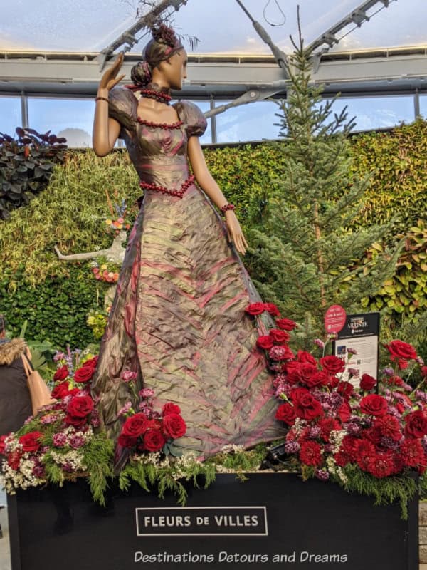 Flora art display features mannequin in dress of red and greenish-brown leaves with fitted bodice and flared skirt surrounded by red roses