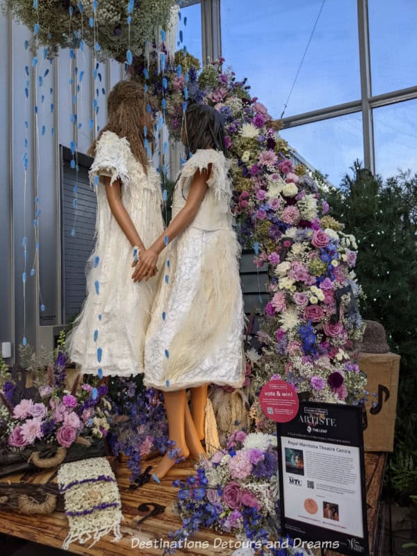 Floral art display shows two female mannequins dressed in white facing each other and framed with a half circle of rose, blue, and white flowers