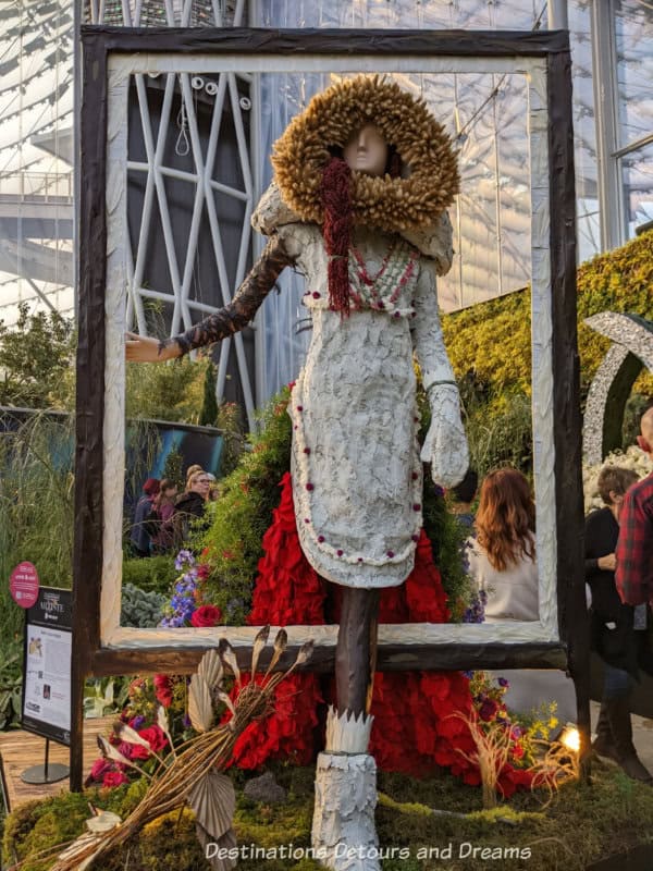 Floral art display features mannequin in white parka dress with hood stepping through a window frame, her train of red flowers behind her