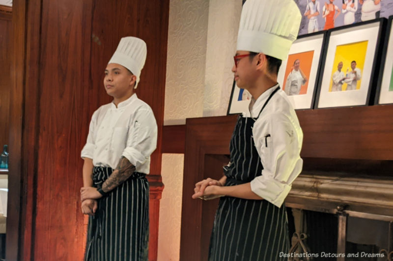 Two chefs in white hats, white shirts, and black aprons with white stripes talking to a dining room