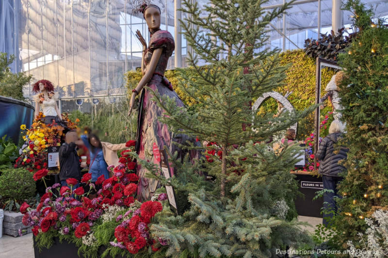 Display room inside an indoor garden featuring mannequins decorated with floral displays, evergreens, and walls on either side containing a vertical wall of plants and a northern lights display