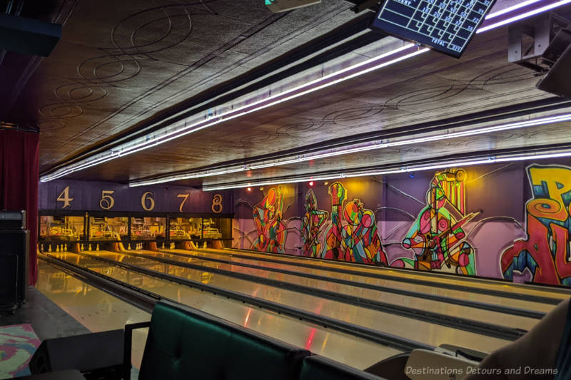 Several bowling lanes with an art deco ceiling and a side wall containing a mural of musical instruments