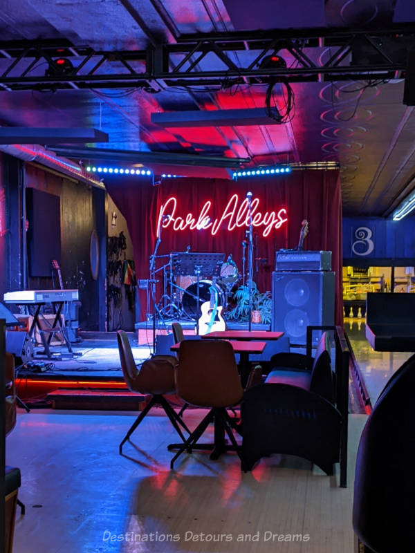 Music venue stage area with drum set in back, keyboard to side, large amp, guitar resting on stand and neon sign in front of red curtain at back that says Park Alleys