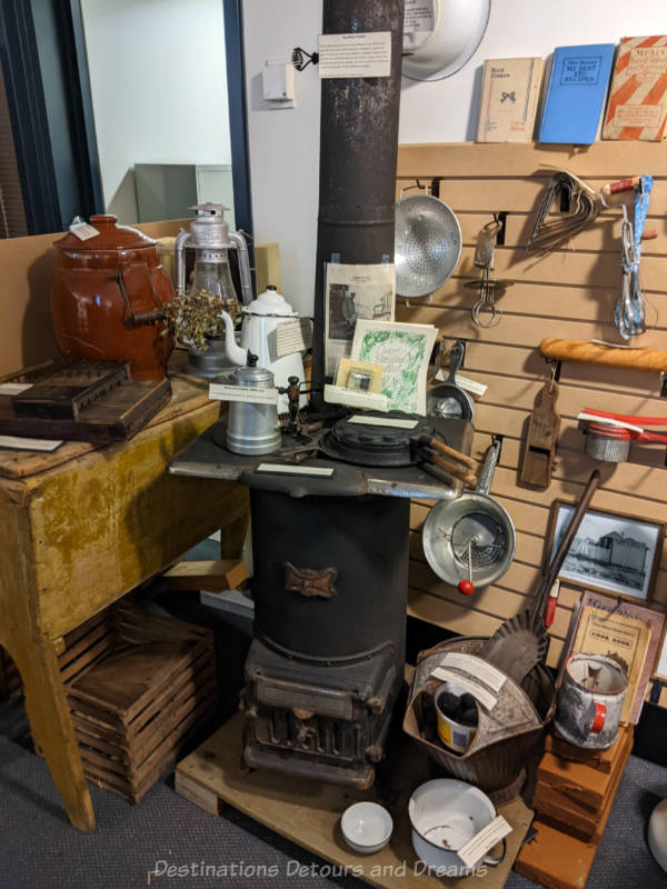 A display of early twentieth century items at a museum around an old wood stove with a dough keeper at the side and a variety of cooking items